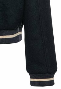 Manche tricotée noire avec un poignet côtelé présentant un motif rayé beige et gris, partie inférieure de l'ourlet d'un vêtement visible.