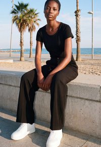 Junge Person in schwarzem Outfit und weißen Sneakers sitzt auf einer Betonbank am Strand mit Palmen und Ozean im Hintergrund.