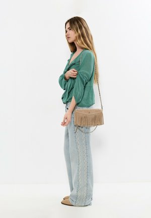 Mujer con blusa verde, jeans anchos con bordados claros, bolso de ante con flecos color tostado y zapatos sin cordones color tostado, de perfil.