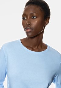 Chemise à manches longues bleu clair avec un col rond, fabriquée à partir d'un tissu doux, présentée sur un fond neutre, mettant en valeur sa texture lisse.