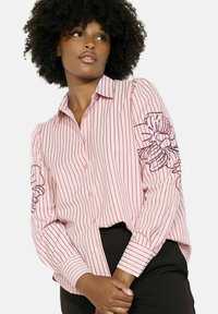 Pruhovaná ružová košeľa s predným zapínaním na gombíky, nafúknutými rukávmi a kvetinovou výšivkou na ľavom rukáve. Vyrobená z ľahkej tkaniny.