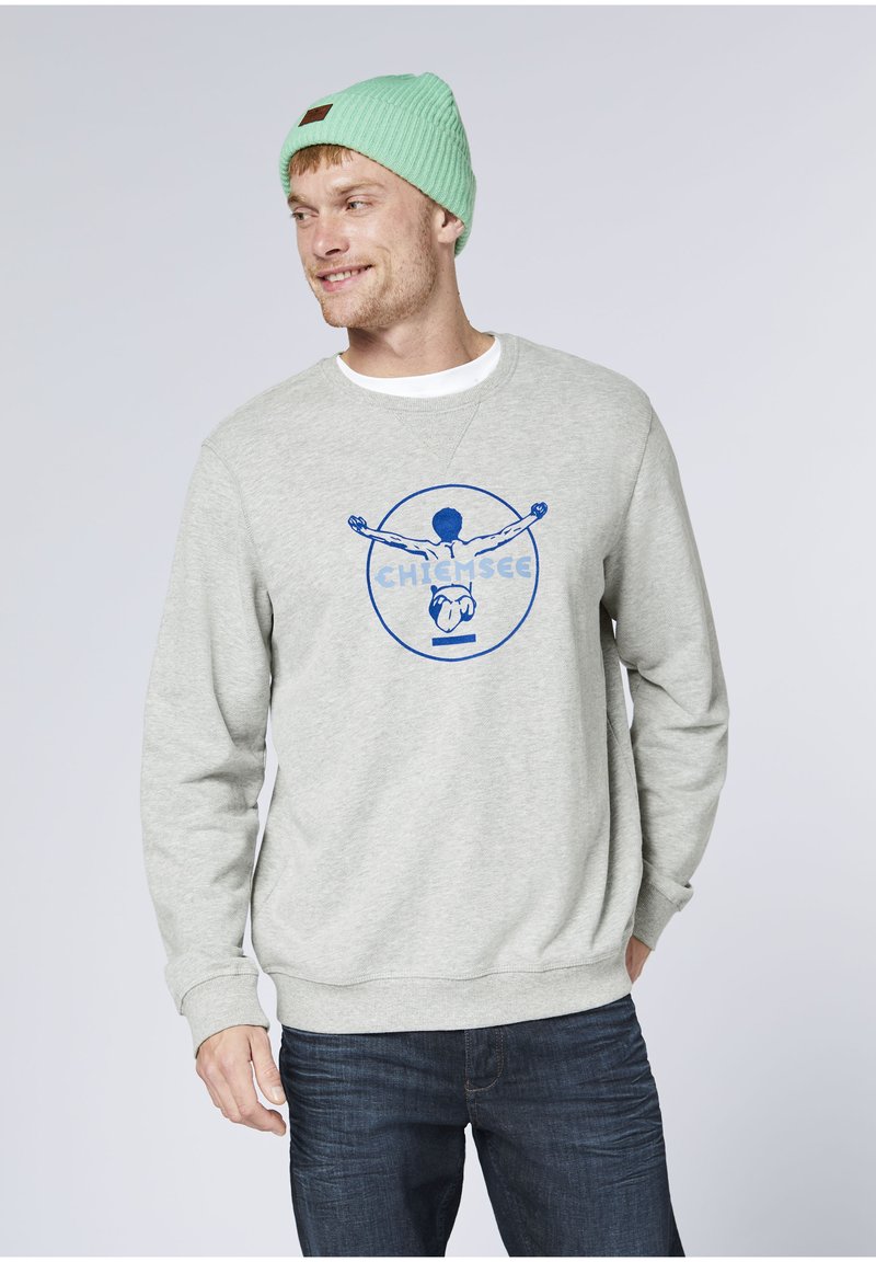 Gray sweatshirt featuring a blue graphic design with a figure and "CHIEM SEE" text. Paired with a light green knit beanie. Dark jeans visible.