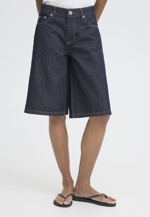 Tmavomodré džínsové šortky po kolená nosené s čiernymi žabkami, zobrazujúce predné vrecká, gombík a zips.