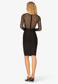 Schwarzes Kleid mit langen, durchsichtigen Mesh-Ärmeln, die mit kleinen Punkten verziert sind, taillierter Silhouette, Reißverschluss auf der Rückseite und knielanger Länge.