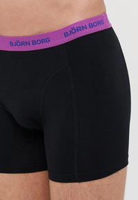 Boxer-briefs noirs avec une ceinture en élastique violette affichant "BJÖRN BORG" en lettres bleues. Tissu lisse avec une coupe ajustée et des coutures plates.
