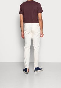 Witte broeken met een rechte pasvorm, gecombineerd met een bruine crewneck t-shirt en blauwe high-top sneakers met bruine accenten. Effen achtergrond.