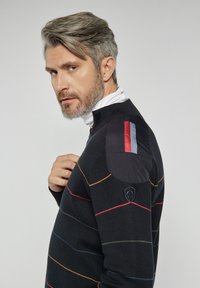Czarny sweter z wielokolorowymi poziomymi paskami. Posiada stójkę, poduszkowe akcenty na ramionach w czarnym kolorze oraz logo w kształcie tarczy.