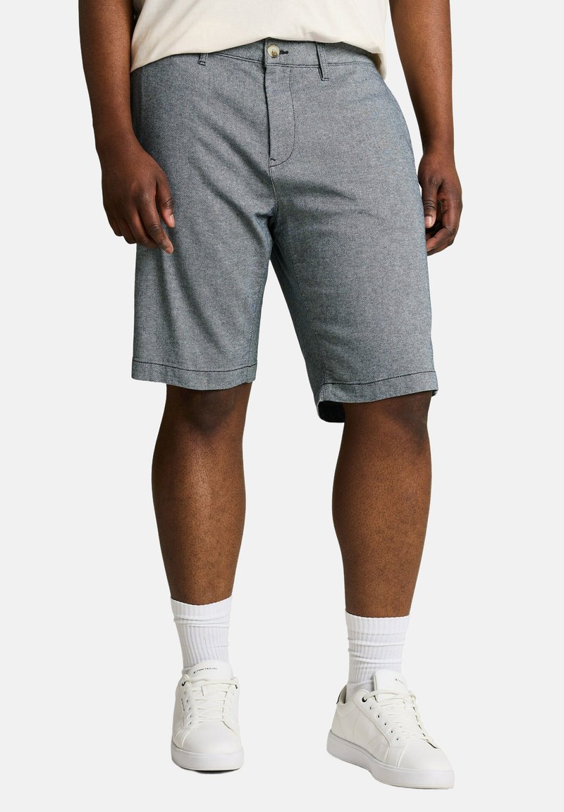 Mann trägt graue knielange Shorts, weiße Sportsocken und weiße Turnschuhe, steht mit entspannten Händen an den Seiten.