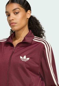 Burgundy zip-up jacket made of smooth fabric, featuring white three-stripe accents on sleeves and a white logo on the left chest.