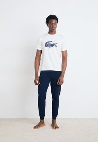 Un tricou alb cu un grafic de crocodil bleumarin, asortat cu pantaloni ajustați bleumarin. Materialul pare moale și are un design casual.