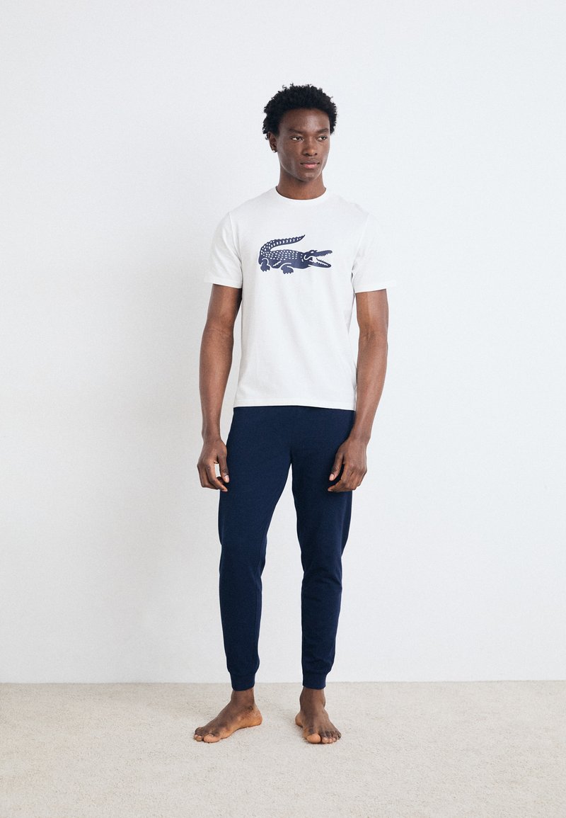 Un tricou alb cu un grafic de crocodil bleumarin, asortat cu pantaloni ajustați bleumarin. Materialul pare moale și are un design casual.