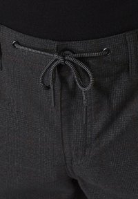 Graue Karohose aus strukturiertem Stoff, mit einem schwarzen Kordelzugverschluss und zwei seitlichen Taschen. Subtile braune Akzente im Muster.