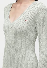 Femme portant un pull en maille gris clair avec motif torsadé et col en V profond, affichant un petit logo mouette rouge sur la poitrine.
