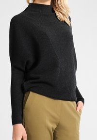Black ribbed sweater with a high neckline, relaxed fit, and diagonal design. Paired with light brown trousers. Textured fabric.