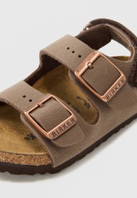 Sandal de couro castanho com duas tiras ajustáveis com fivelas em tom cobre, palmilha em cortiça moldada e sola de borracha texturizada.