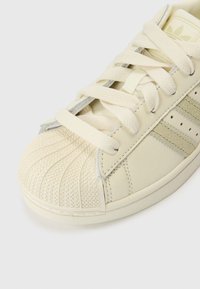 Ténis de couro creme com biqueira de borracha texturizada, atacadores planos e riscas de realce tricolores. Apresenta detalhes costurados e perfurações.