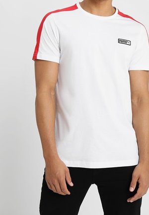 Mann in einem weißen Puma-T-Shirt mit roten Schulterstreifen und schwarzen Hosen, der vor einem schlichten hellen Hintergrund steht.