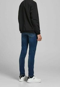 Svart bomberjacka, blå denimjeans och vita sneakers. Bakifrån visar den texturerade tyget av jackan, slim-fit jeansen och den distinkta skodesignen.