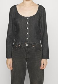 Veste en denim noir raccourcie avec des manches longues, un col rond et une fermeture à boutons argentés sur le devant. Porte avec un pantalon en denim foncé.