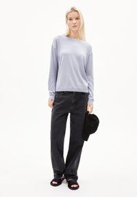 Hellblauer, langärmeliger Pullover in lockerer Passform, kombiniert mit hochgeschnittenen, dunklen Jeans. Eine schwarze Kappe wird in einer Hand gehalten, flache schwarze Sandalen schmücken die Füße.