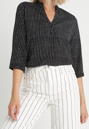 Vrouw draagt een zwarte blouse met verticale witte strepen en witte broek met dunne verticale zwarte strepen, staand tegen een effen achtergrond.