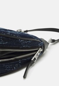 Blå denim crossbody-väska med silverdragkedja, svarta läderdetaljer och justerbar rem; har en texturerad yta och vinkelaktig design.