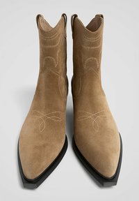Bottes chelsea en daim marron avec bouts pointus, couture décorative et semelle en caoutchouc noire. Conçues avec des anses latérales pour un enfilage facile.