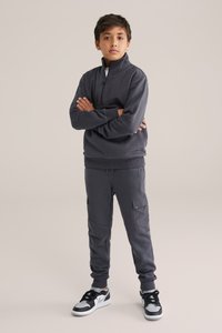 Hanorac zip-up de culoare gri închis, cu guler înalt, asortat cu pantaloni cargo care au buzunare laterale și manșete elastice, purtați cu pantofi sport negri.