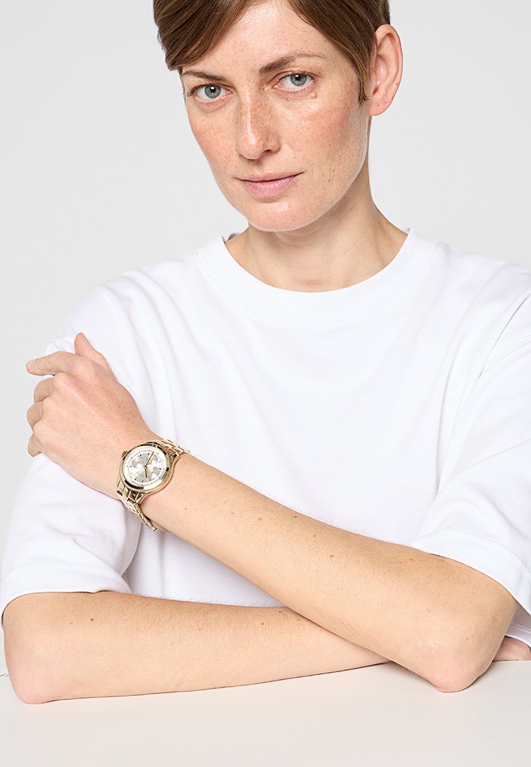 Goldene Uhr mit rundem Ziffernblatt, silbernem Ziffernblatt und schwarzen Akzenten. Am Handgelenk getragen, kombiniert mit einem schlichten weißen Kurzarmhemd. Einfaches, modernes Design.