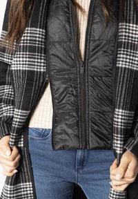 Černá péřová vesta se zipem, vrstvená pod kostkovaným kardiganem v černé a bílé barvě. Obsahuje dlouhé rukávy a strukturovanou tkaninu.