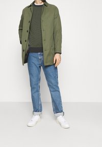 Oliivinvihreä takki napillaisella suljennalla, teksturoitu tummanvihreä neule, vaaleansiniset käärityt farkut ja valkoiset tennarit. Rentoon tyyliin sopiva asu.