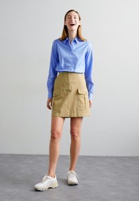 Camicia azzurro chiaro con bottoni, gonna cargo beige con tasche frontali e sneakers beige. Tessuto liscio, design strutturato e colori neutri.