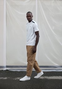 Bijela majica kratkih rukava s crnim detaljima, smeđe cargo hlače s džepovima i bijele sportske tenisice. Model stoji na pločniku.