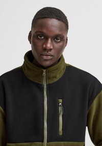 Chaqueta de forro polar con un alto cuello, que presenta paneles en negro y verde oliva, cierres de cremallera y un pequeño bolsillo con cremallera en el pecho.