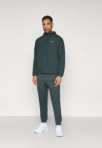 Temnozelena lahka jakna s kapuco, z zadrgo in belim logotipom Nike, v kombinaciji s ustreznimi ozkimi hlačami in belimi supergami.