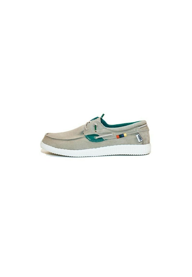 Zapato de lona gris claro con detalles en verde, punta redonda, suela blanca y pequeño detalle de bandera en el lado. Diseño con cordones y perfil bajo.