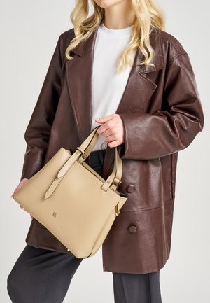 Bolso de mano de cuero beige con doble asa superior, superficie texturizada y detalles en hardware dorado. Combinado con un abrigo de cuero marrón.