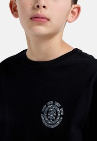 Černé bavlněné tričko s kulatým výstřihem a texturovaným grafickým logem v šedé barvě, obsahující vzory hor v oblasti levého prsa.