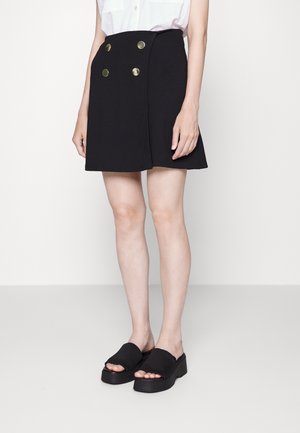 Kobieta ubrana w czarną spódnicę w kształcie litery A z czterema złotymi guzikami oraz czarne sandały na platformie, stojąca na białym tle.