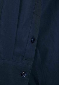 Camicia navy con bottoni, realizzata in un tessuto liscio, dotata di colletto arrotondato, cuciture visibili e un singolo bottone sul polsino.