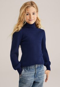 Navy kék bordázott garbós pulóver hosszú ujjakkal, testhezálló fazonnal és puha tapintással, világoskék farmernadrággal párosítva.