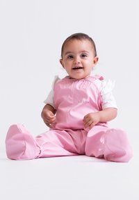 Rosa wasserdichte Latzhose mit Reißverschluss, breiten Beinen und weicher Textur. Darunter ein weißes Kurzarmhemd.