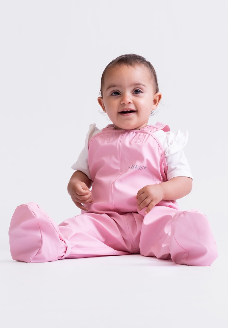 Rosa wasserdichte Latzhose mit Reißverschluss, breiten Beinen und weicher Textur. Darunter ein weißes Kurzarmhemd.