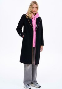 Blondína v čiernom dlhom kabáte s oranžovým a ružovým lemovaním, ružovej mikine, šedých širokých rifle a bielych teniskách stojí s jednou rukou v vrecku.