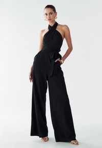 Combinaison noire sans manches avec jambes larges, tissu texturé et détail de taille nouée. Le mannequin porte des boucles d'oreilles en métal doré et des sandales.