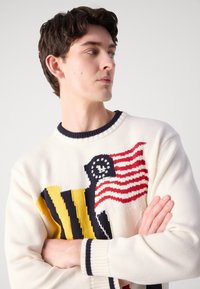 Pull en tricot crème avec un col rond bleu marine, présentant un design graphique vibrant de rayures et de drapeaux en rouge, jaune et bleu marine.