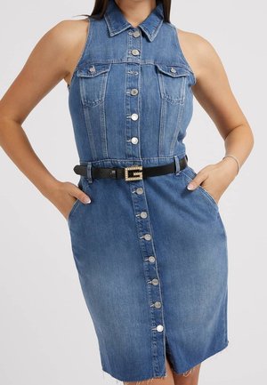 Niebieska sukienka bez rękawów z denimu z guzikami z przodu, dwoma kieszeniami na piersi, czarnym paskiem z kwadratową klamrą i rękami w kieszeniach bocznych.