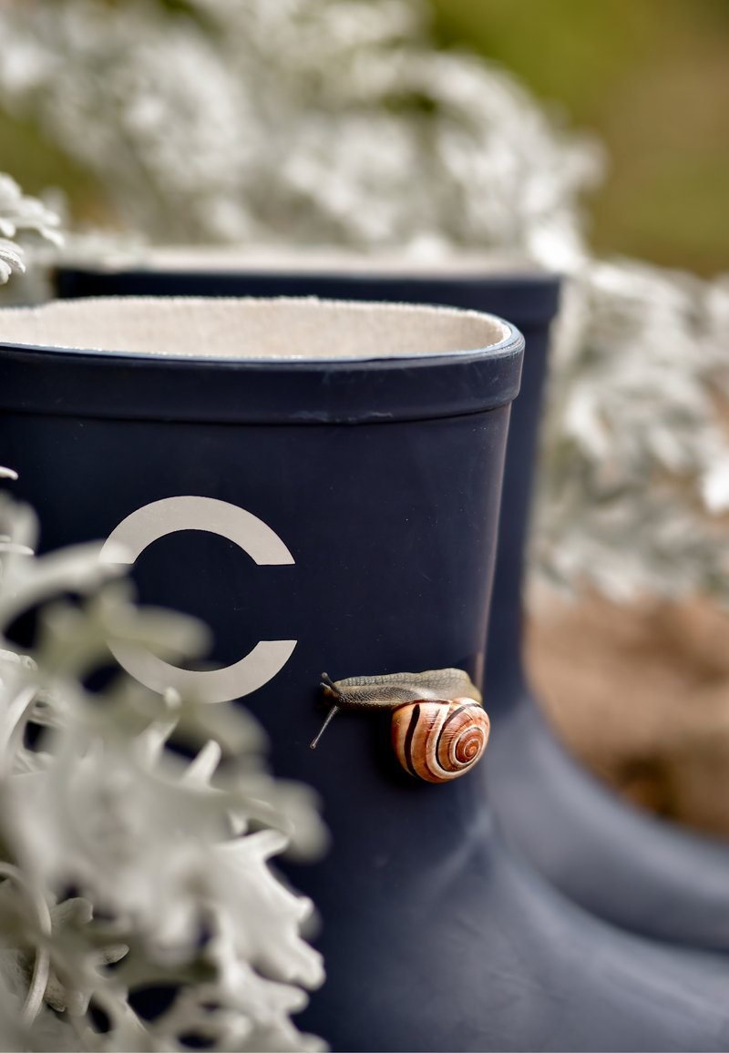 Tengerészkék gumicsizma fehér kör alakú logóval; egy barna csíkozású csiga pihen az oldalán, zöld növényzet veszi körül.