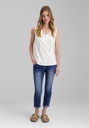 Junge Frau mit langen blonden Haaren, die ein weißes ärmelloses Top, hochgekrempelte blaue Jeans und beige Sandalen trägt, steht vor einem grauen Hintergrund.
