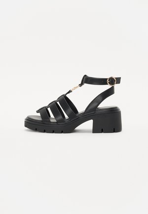 Sandalias de tacón bloque de cuero negro con múltiples correas anchas, una correa ajustable en el tobillo y acentos de hardware en tono dorado. Suela de goma plana.